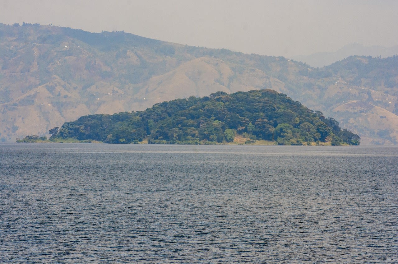 An island covered in lush greenery sits in the middle of a serene lake with mountainous terrain in the background.